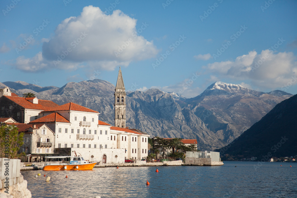 Amazing old town with tower by sea, snow mountains, clouds. Trip to Perast with tiled roof, palms, boats by the Adriatic Sea in Montenegro.