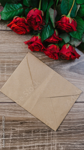 Bouquet of red roses on a wooden table with an envelope and a blank paper card for your text