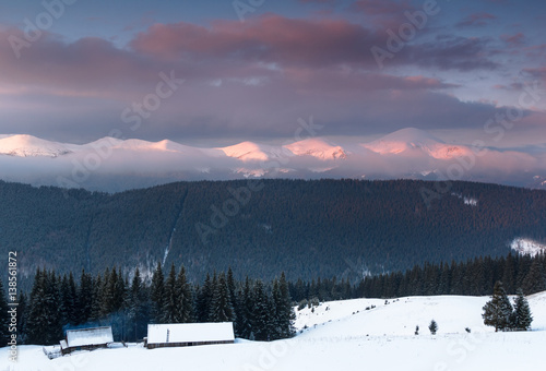 Landscape of the old farm in the winter mountains at sunrise. Dramatic overcast sky and distance view on snow capped peaks.