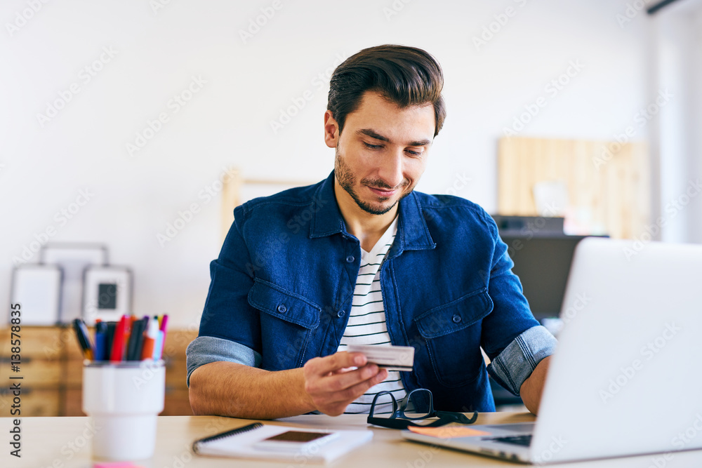 Fototapeta premium Young man doing online payments typing number from his credit card to laptop computer