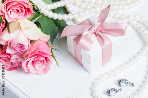 Present box with pink ribbon with jewellery and roses on white table