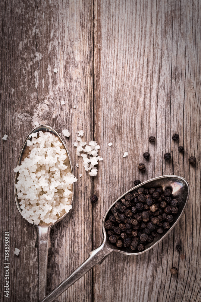 Salt crystals and black peppercorns in old silver spoons. Salt crystals and black peppercorns in old silver spoons.