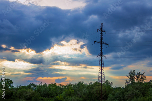high-voltage tower against the dark dramatic clouds through which the sun's rays make their way