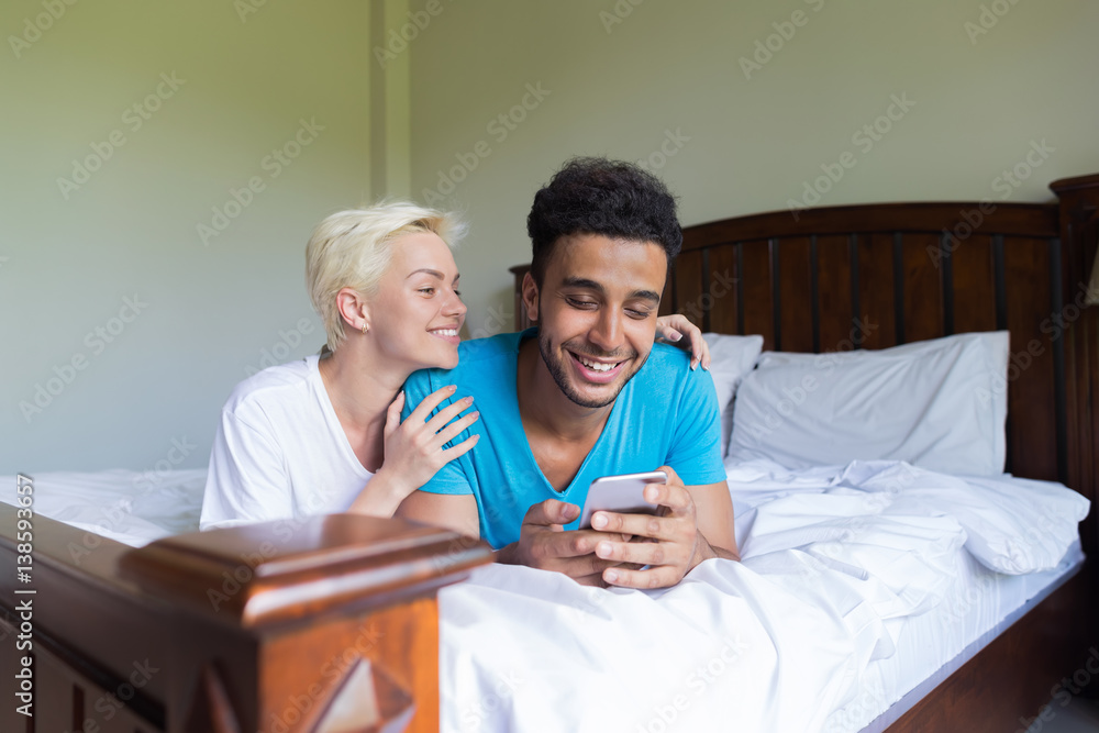 Young Couple Lying On Bed, Happy Smile Hispanic Man And Woman Using Cell Smart Phone , Lovers In Bedroom Young Couple Lying On Bed, Happy Smile Hispanic Man And Woman Using Cell Smart Phone , Lovers In Bedroom
