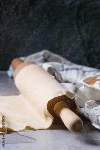 Fresh homemade rolled dough for pasta tagliatelle on wood rolling pin with egg yolk and kitchen towel over dark gray table background.