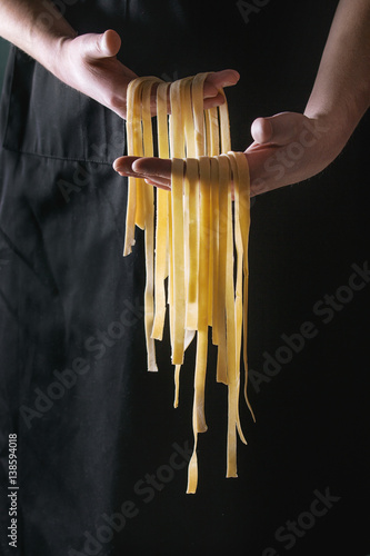 Fresh raw uncooked homemade pasta tagliatelle in man's hands over black apron as background.