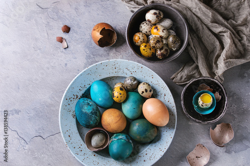 Colored Easter blue brown chicken and quail eggs, whole and broken with yolk in shell in spotted plate and black bowls with textile over gray textured background. Top view with space