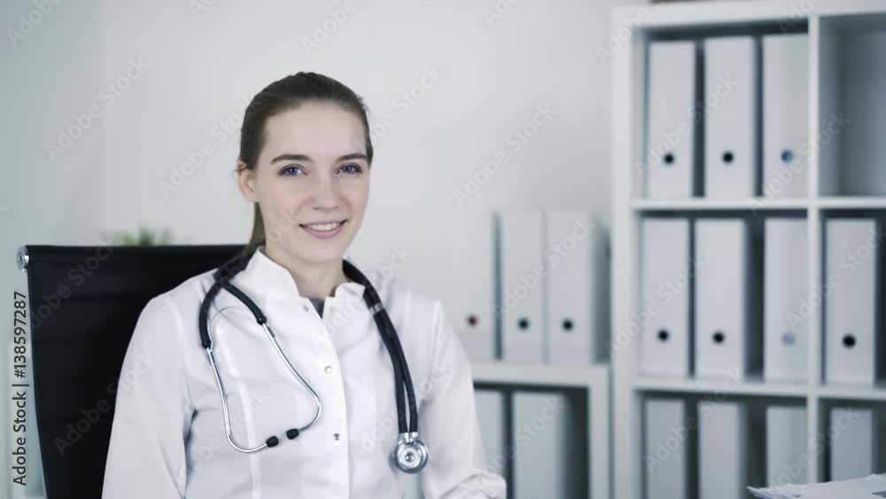 Young beautiful general practicer is twisting on the chair at medical office, looking at camera and smiling. Locked down real time portrait shot. Young beautiful general practicer is twisting on the chair at medical office, looking at camera and smiling. Locked down real time portrait shot.