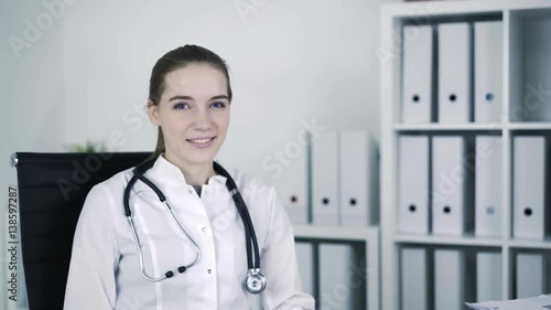 Young beautiful general practicer is twisting on the chair at medical office, looking at camera and smiling. Locked down real time portrait shot. photo