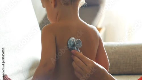 Kid doctor examing little baby boy with stethoscope in a sunny room
