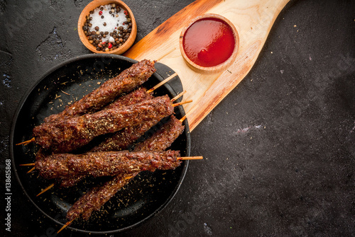 Shish kebab on a stick, from ground beef meat. Lula kebab, traditional Caucasian dish. On the black background of the concrete, on a chopping board, with ketchup, spices and tomatoes copy space above