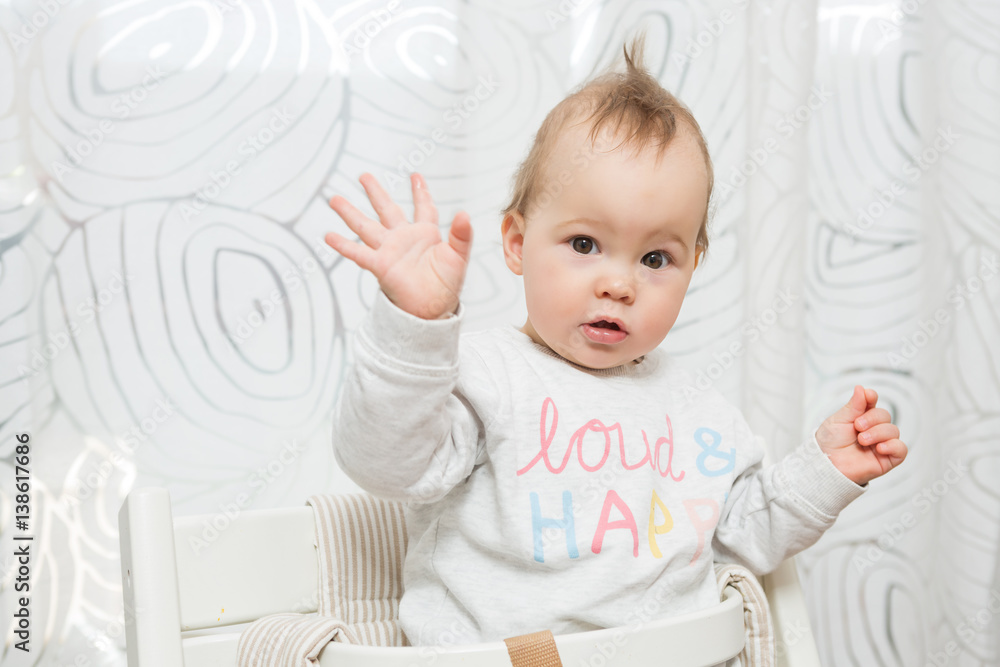 Eleven months old baby girl sitting in a stool making funny faces into the camera, raising her both hands in the air Eleven months old baby girl sitting in a stool making funny faces into the camera, raising her both hands in the air