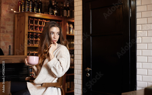 Charming European girl sitting at the bar with a big cup of cappuccino with his finger to his mouth attached on the background wine cabinet and looking away thoughtfully. Copy space for your content