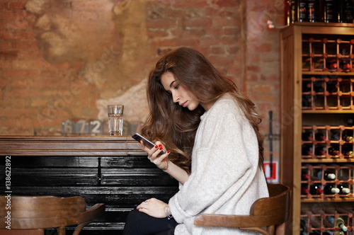 Handsome Caucasian young woman using free wi-fi on mobile phone, sitting at the bar on the background of an old brick wall, looking at the screen with serious and concentrated expression