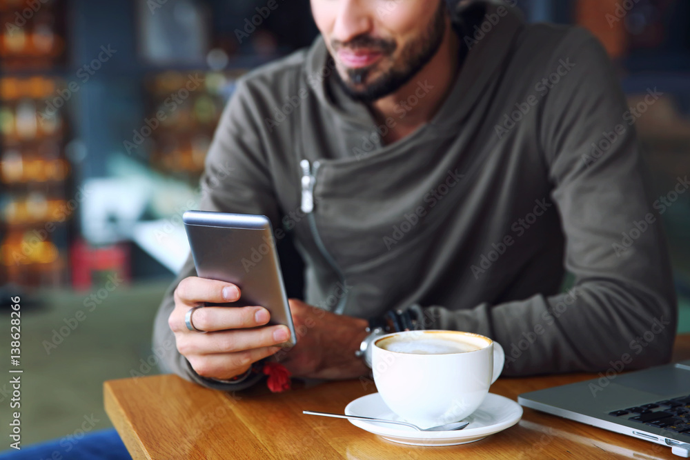 Young handsome cheerful hipster guy at the restaurant using a mobile phone, hands close up. Selective focus. Young handsome cheerful hipster guy at the restaurant using a mobile phone, hands close up. Selective focus.
