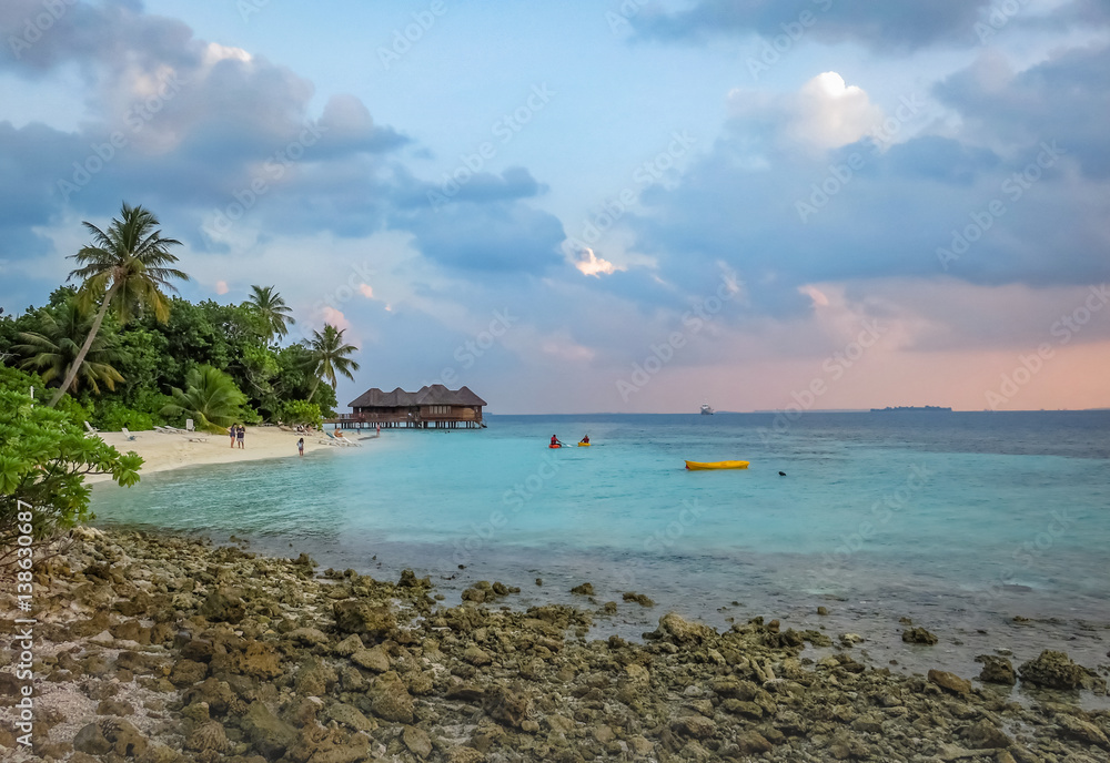 Landscape of a beautiful tropical rocky and sandy beach in Maldives island. Blue ocean water and cloudy, sunset sky in the background. Palms, wooden bungalow, villa, people kayaking.