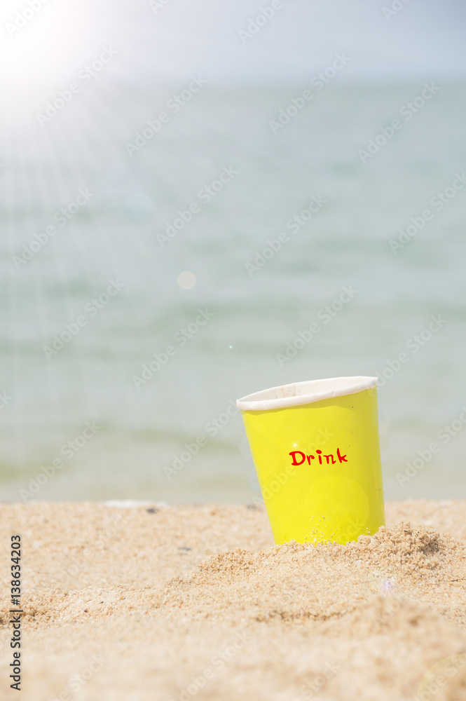 Yellow paper glass beverage on the sand behind beach with sunlight.