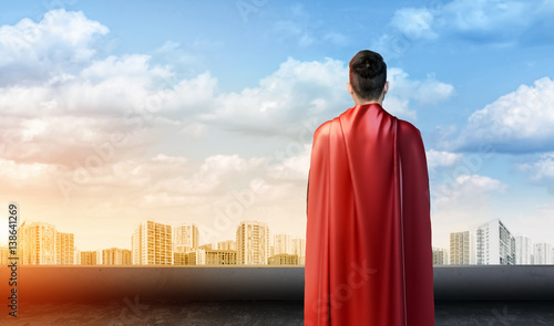 A businessman in superman cape standing turned back on the sky background with cityscape below.