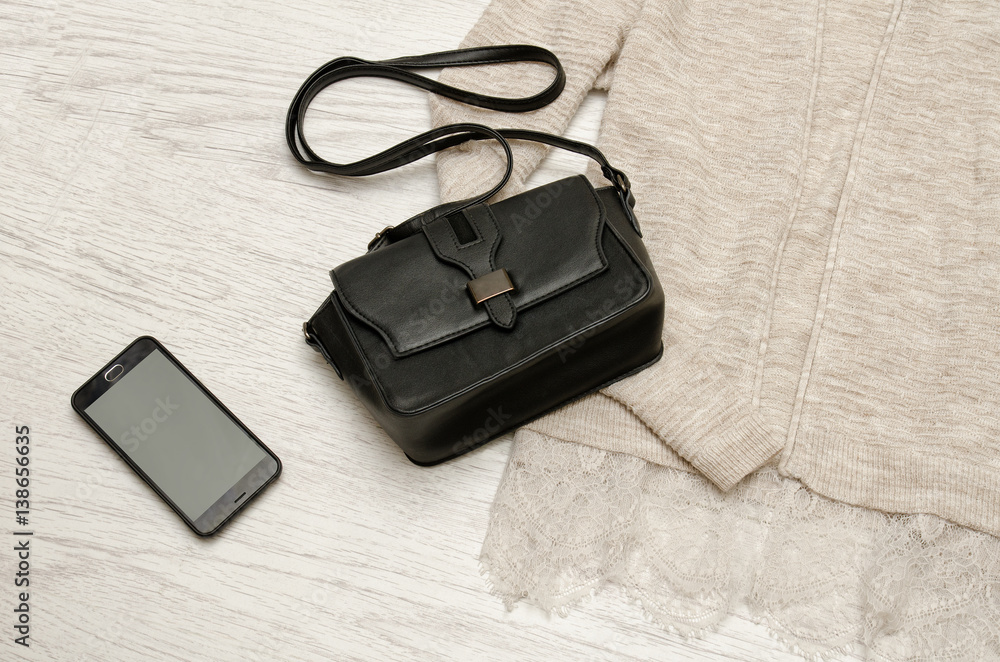 Black handbag and mobile phone on a beige cardigan. Wooden background. Fashionable concept, top view Black handbag and mobile phone on a beige cardigan. Wooden background. Fashionable concept, top view