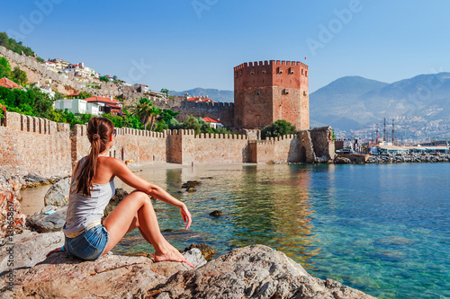 Young woman look at Kizil Kule tower in Alanya peninsula, Antalya district, Turkey, Asia. Famous tourist destination with high mountains. Part of ancient old Castle. Summer bright day photo