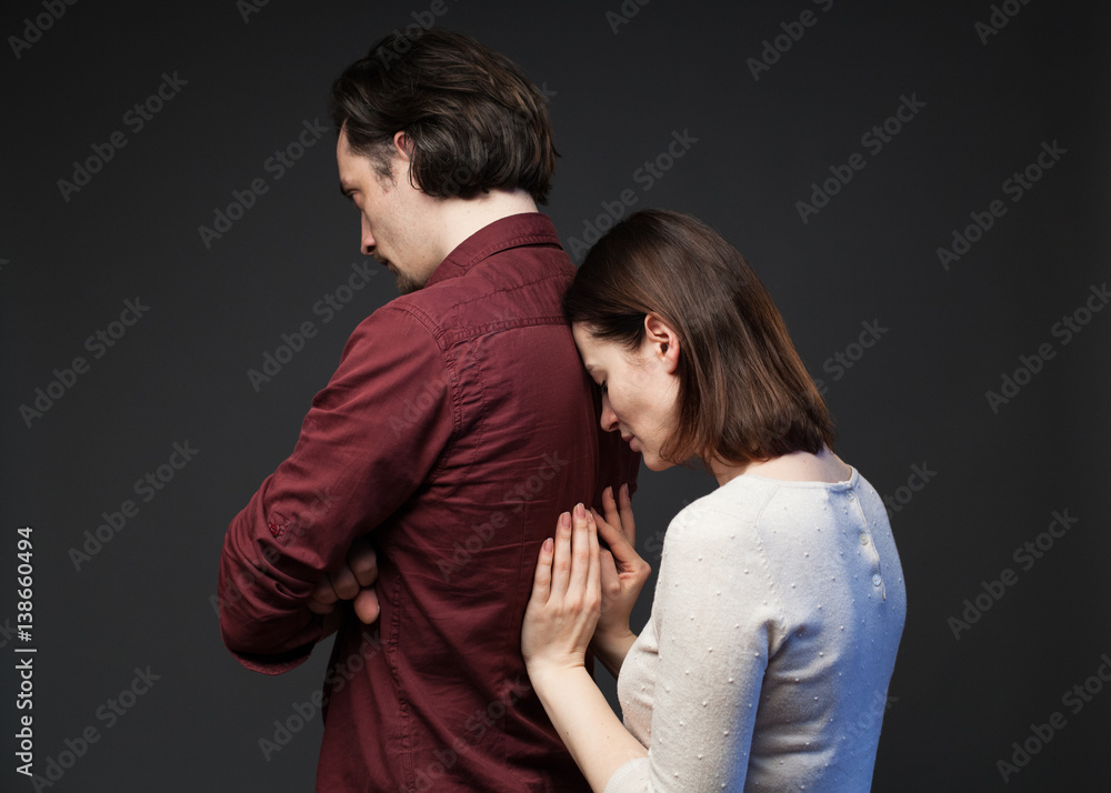 Misunderstanding in the family - the man is standing turned away with crossed hands, the woman is touching his back tenderly, gray background Misunderstanding in the family - the man is standing turned away with crossed hands, the woman is touching his back tenderly, gray background