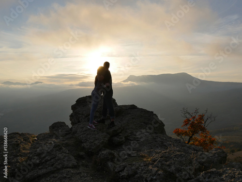Silhouettes of young couple standing on a mountain and looking to each other on beautiful sunset background. Love of guy and girl.