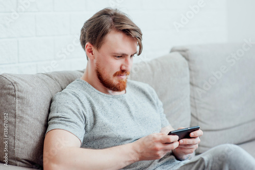 The bearded man sitting on the couch at home and playing the phone