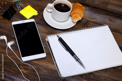 White mobile phone on wooden wooden background with headphones  cup of coffee  croissant and Stationery appliances