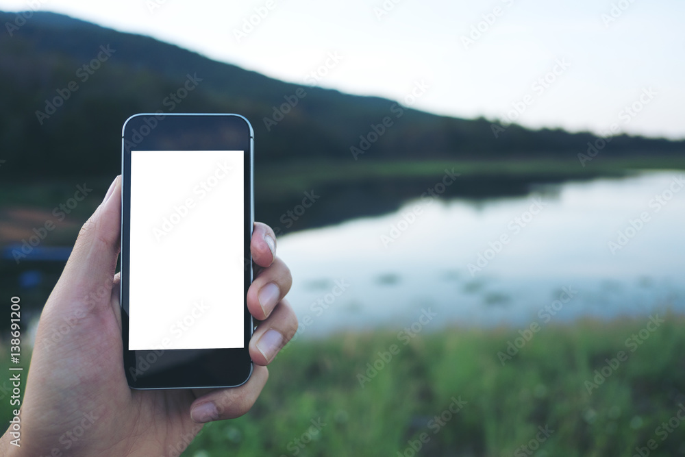 Mockup image of hand holding black mobile phone with white blank screen at outdoor and lake nature background