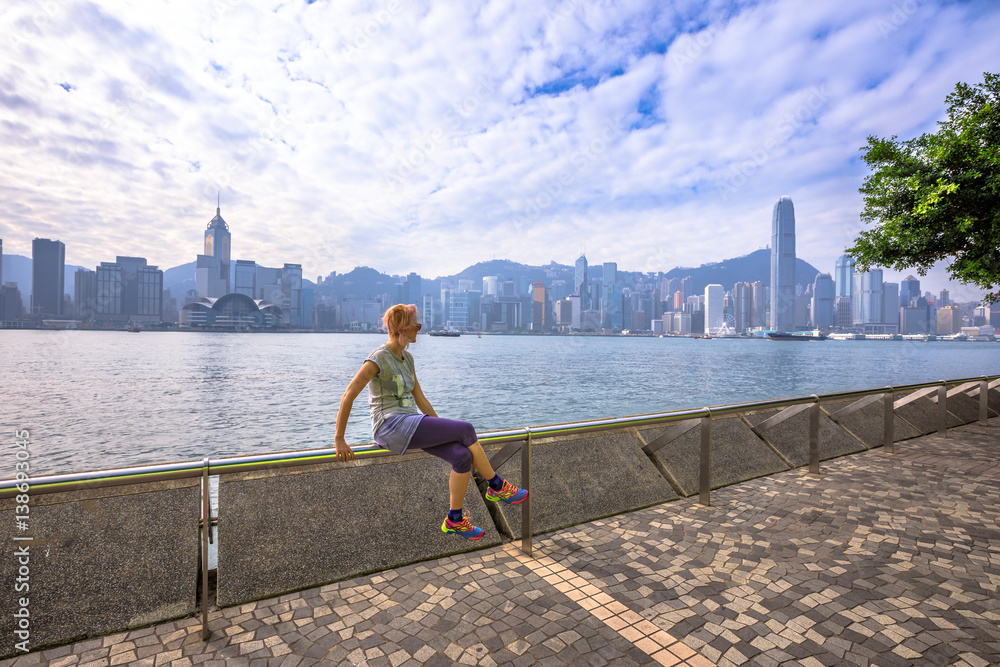 Naklejka premium Caucasian woman with urban background Hong Kong skyline. Female fitness athlete after a workout on Tsim Sha Tsui Promenade and Avenue of Stars in Victoria Harbour, Kowloon. Healthy lifestyle concept.