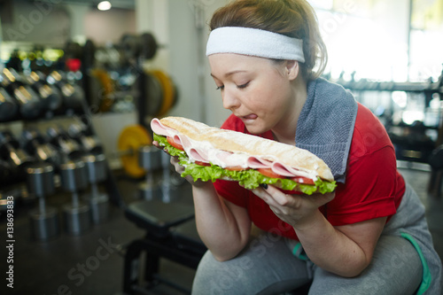 Portrait of young overweight woman licking her lips wanting to eat huge fat sandwich while working out in gym  struggling to keep fit