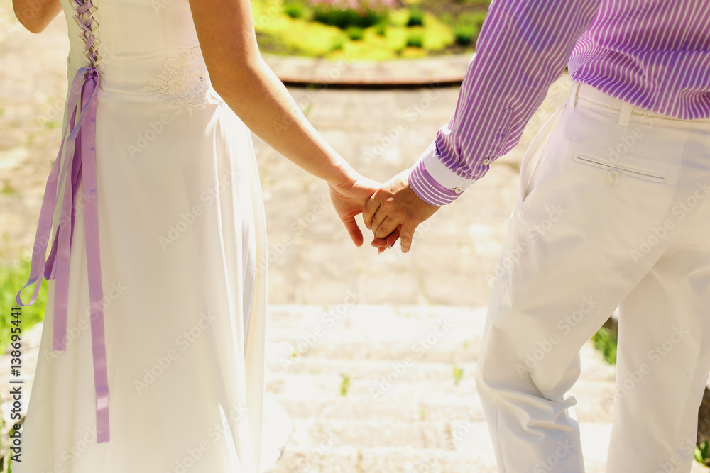 Groom in violet shirt and bride in dress with violet ribbons hold each other hands while walking