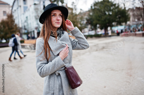Young model tourist girl in a gray coat and black hat with leather handbag on shoulders posed at street of city.