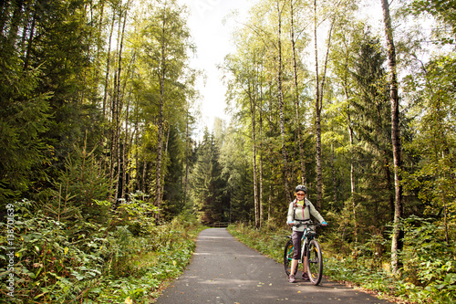 Female tourist cyclist standing on road in forest, looking to camera and smiling.