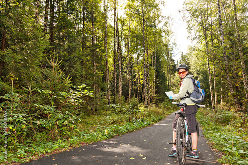 Female tourist cyclist standing on road in forest with map, looking to camera and smiling.