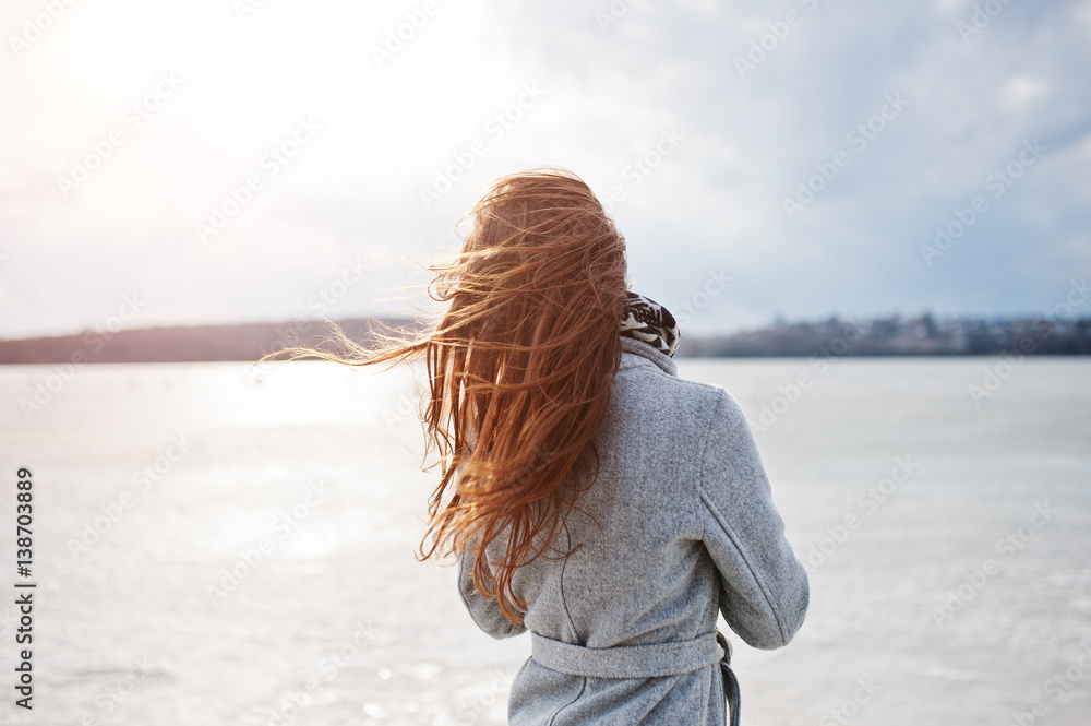 Back of young model girl in gray coat with red hair against freeze lake. Back of young model girl in gray coat with red hair against freeze lake.