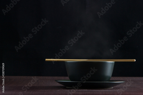 Black bowl with smoke boiling up and wooden chopsticks on wooden table with black background