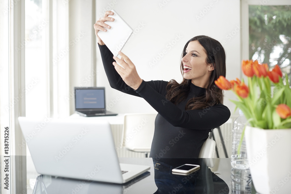 Capture the moment. Shot of a young businesswoman sitting in front of laptop and taking selfie with her digital tablet phone while working from home. Capture the moment. Shot of a young businesswoman sitting in front of laptop and taking selfie with her digital tablet phone while working from home.