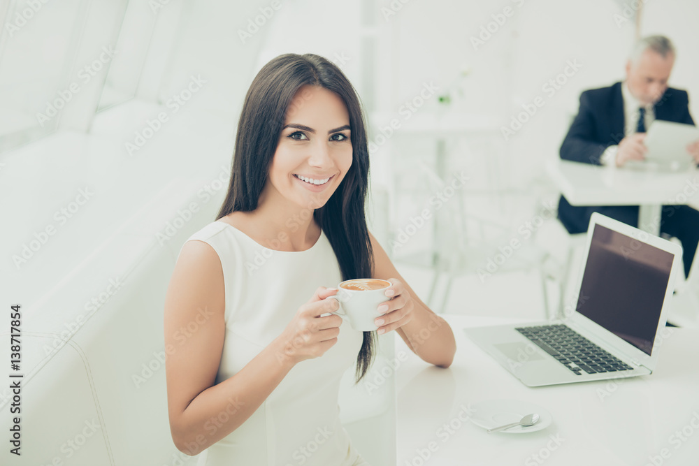 A portrait of happy smiling woman drinking a cup of hot coffee and sitting at the table with a laptop A portrait of happy smiling woman drinking a cup of hot coffee and sitting at the table with a laptop