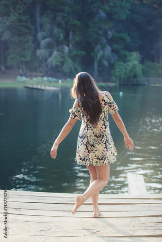 Fototapeta Naklejka Na Ścianę i Meble -  Back view of lady walking on wooden pier near the pond. Vertical outdoors shot.