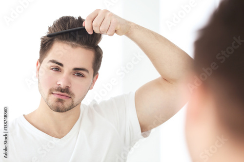 Handsome young man combing hair while standing in front of mirror