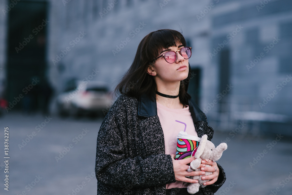 portrait of a beautiful and stylish girl in glasses holding a coffee and a soft toy on the background of the city