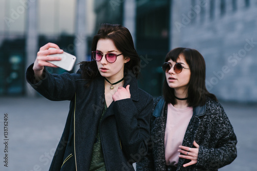 Portrait of two stylishly dressed girls doing selfie against the background of a beautiful cityscape