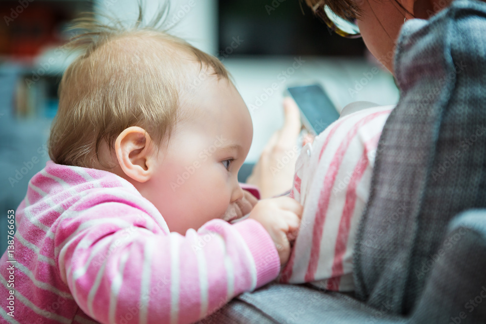 Ten months old baby girl trying to get her mother's attention; mother is sitting in a sofa texting messages on her smartphone Ten months old baby girl trying to get her mother's attention; mother is sitting in a sofa texting messages on her smartphone
