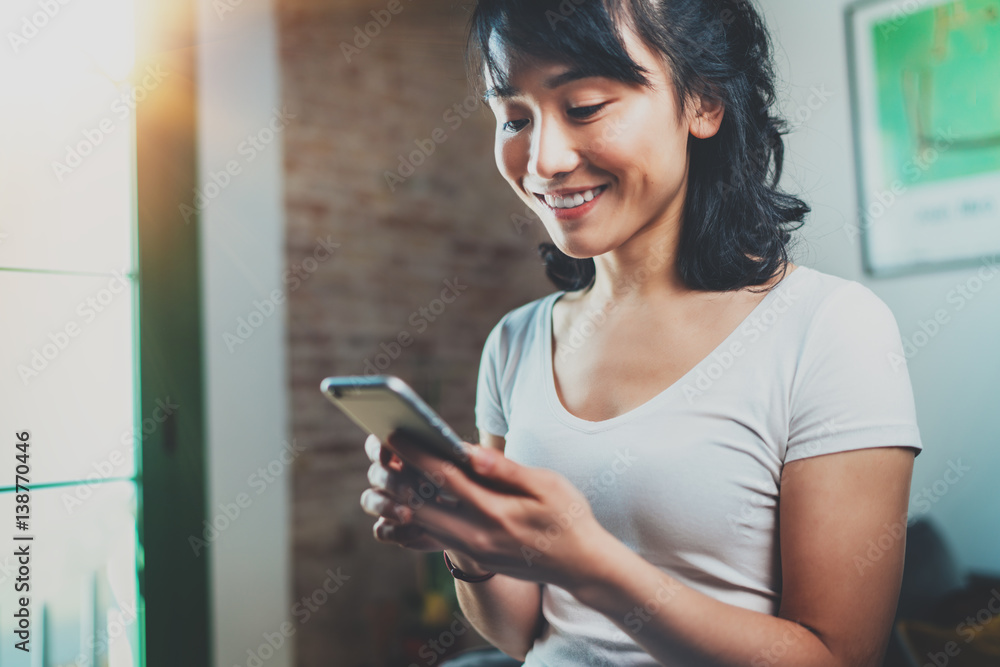 Happy young Asian woman holding mobile phone on hands and reading message at home room interior on the background. Flares. Happy young Asian woman holding mobile phone on hands and reading message at home room interior on the background. Flares.