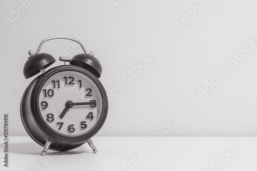 Closeup alarm clock for decorate show a quarter past seven or 7:15 a.m.on white wood desk and cream wallpaper textured background in black and white tone with copy space