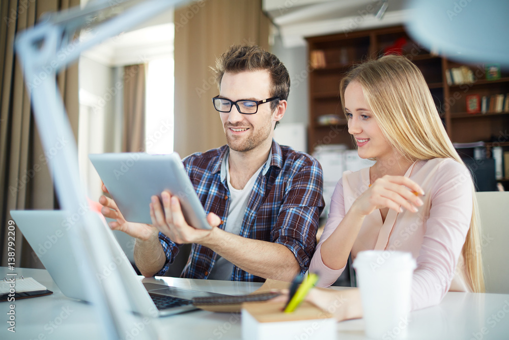 Scene of work process in office: handsome creative man dressed in casual clothes proudly holding up tablet in his hands showing something to blond woman and looking at the screen with curiosity Scene of work process in office: handsome creative man dressed in casual clothes proudly holding up tablet in his hands showing something to blond woman and looking at the screen with curiosity