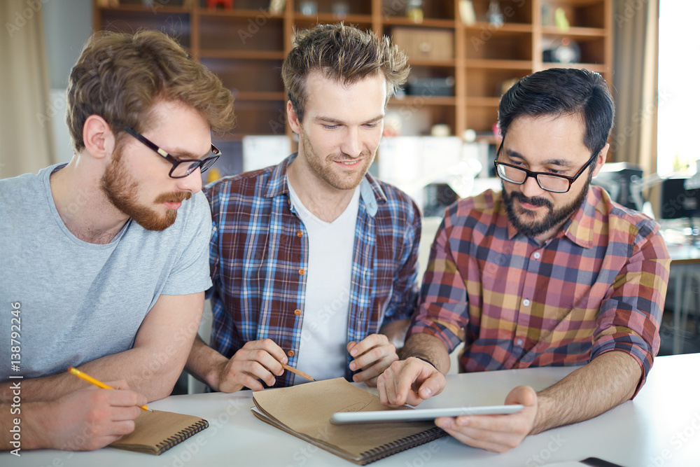 Team of young entrepreneurs at work: three young ambitious men sitting at table planning strategy to new business idea, Asian man pointing at tablet screen, the other two ready to make notes Team of young entrepreneurs at work: three young ambitious men sitting at table planning strategy to new business idea, Asian man pointing at tablet screen, the other two ready to make notes