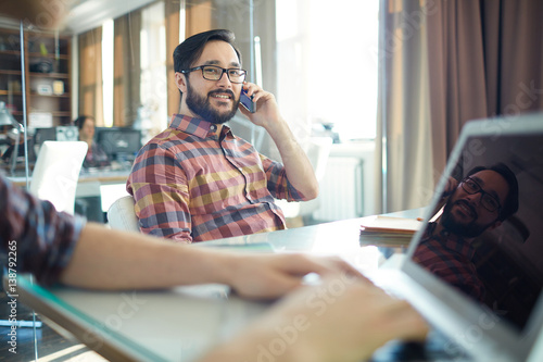 Successful Asian businessman dressed in casual checkered shirt, sitting back in chair in light spacious office and talking on phone, while his colleague working with laptop