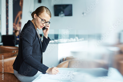 Blond confident businesswoman going over details of contract talking with partner on phone during her coffee break in modern light lounge area